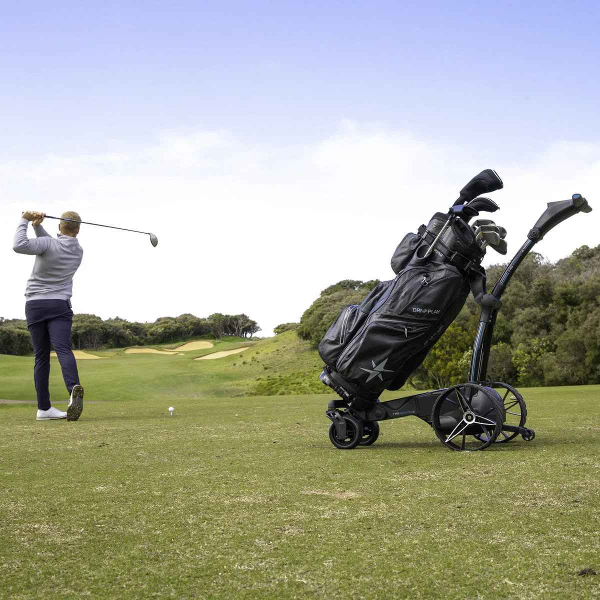 Electric remote golf buggy with golf bag on fairway, golfer teeing off in background