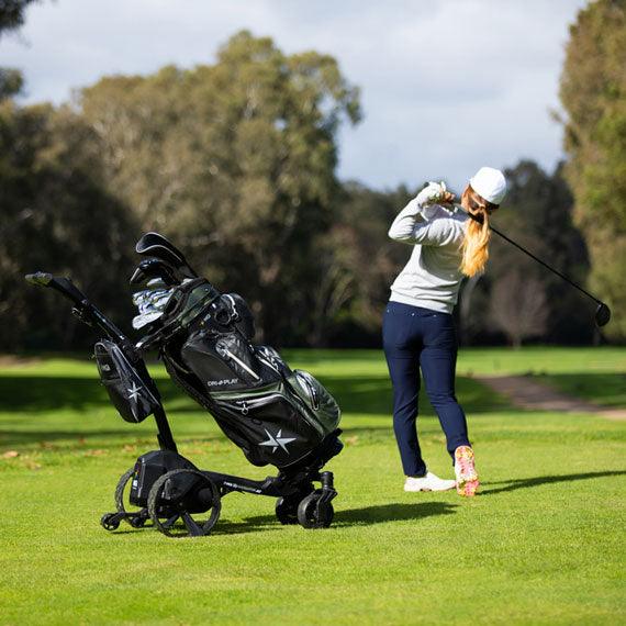 Golfer swinging club on course beside electric golf buggy with golf bag, Hillside Buggies